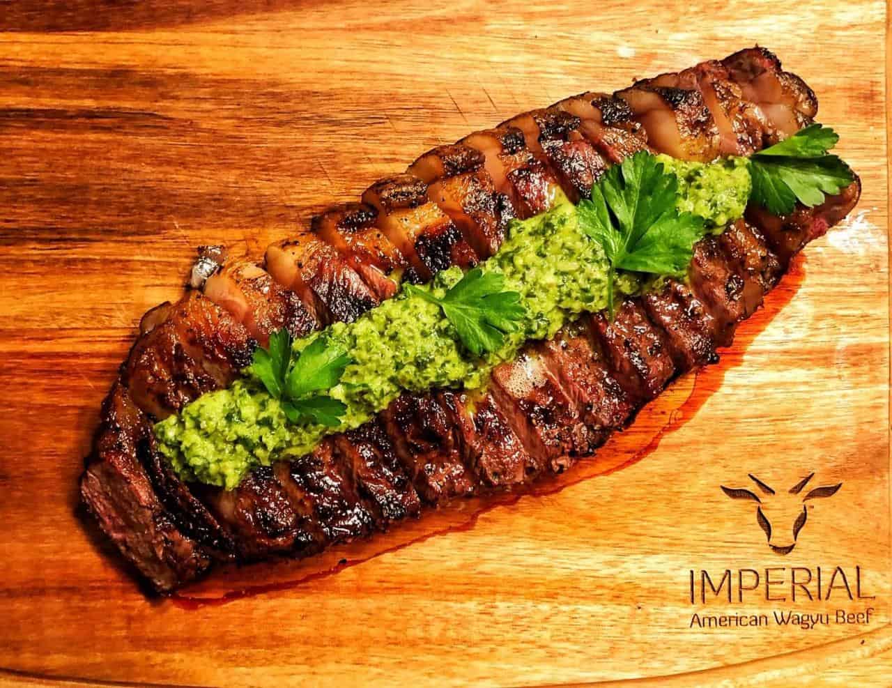 Grilled and sliced Imperial American Wagyu skirt steak topped with chimichurri on a wood board